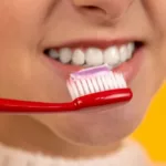 a girl who smiles and brushes her teeth with a red toothbrush on a yellow background